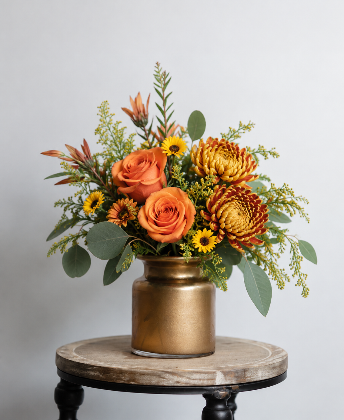 Golden Harvest fall floral arrangement featuring orange roses, bronze chrysanthemums, yellow asters, eucalyptus, and grevillea arranged in a warm golden-bronze vase.
