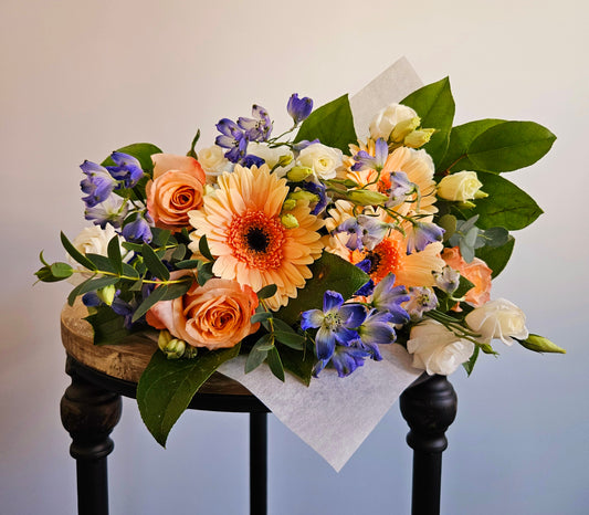 Peach Sunset and Indigo Skies wrapped bouquet peach, blue and white flowers artfully wrapped in parchment paper and tied with a ribbon.