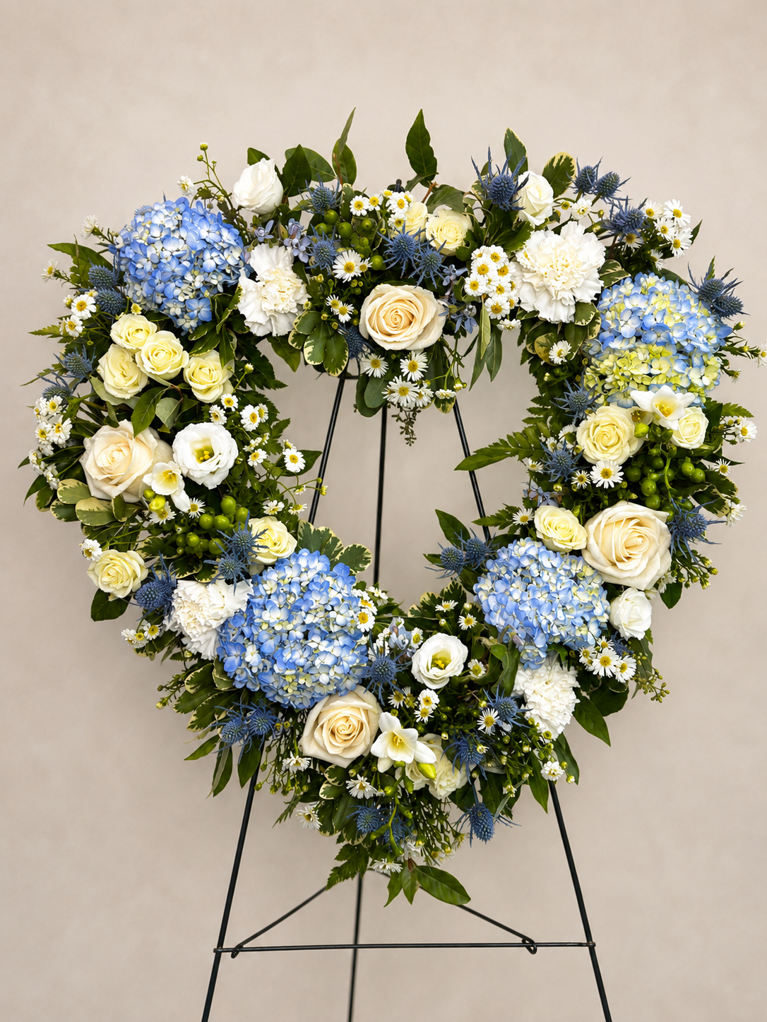 A large floral wreath on a black metal display stand, featuring blue hydrangeas, cream roses, white daisies, and blue thistles against a beige background.