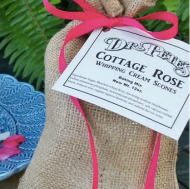 Burlap bag with a pink ribbon and a label for 'Dr. Pete's Cottage Rose Whipping Cream Scones' on a green leafy background.