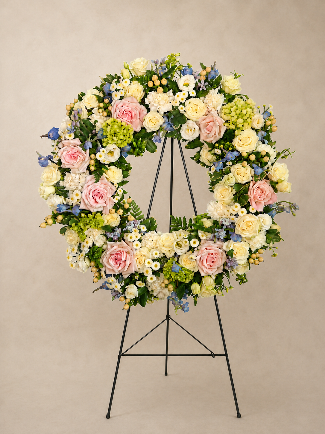 Graceful Remembrance pastel funeral wreath with soft pink roses, ivory blooms, blue accents, green hydrangea and lush greenery displayed on an easel.