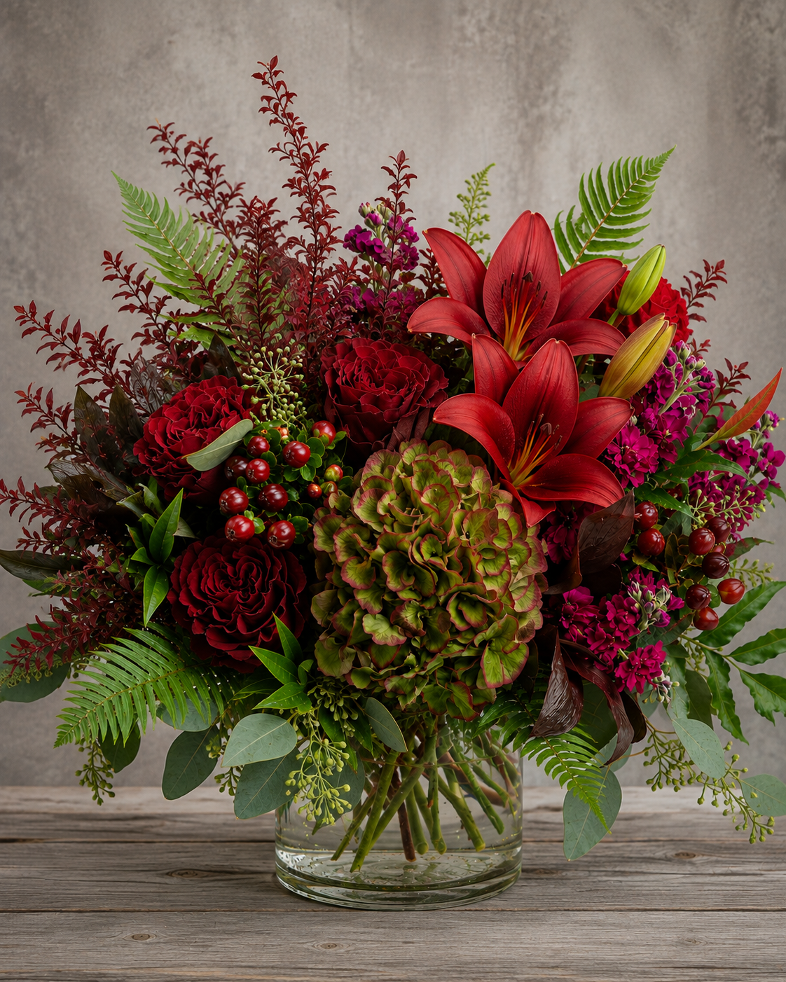 October Meadow fall floral arrangement with red roses, Antique green hydrangea, red lilies, burgundy stock flower, burgundy hypericum berries and textured greenery in a clear glass vase.