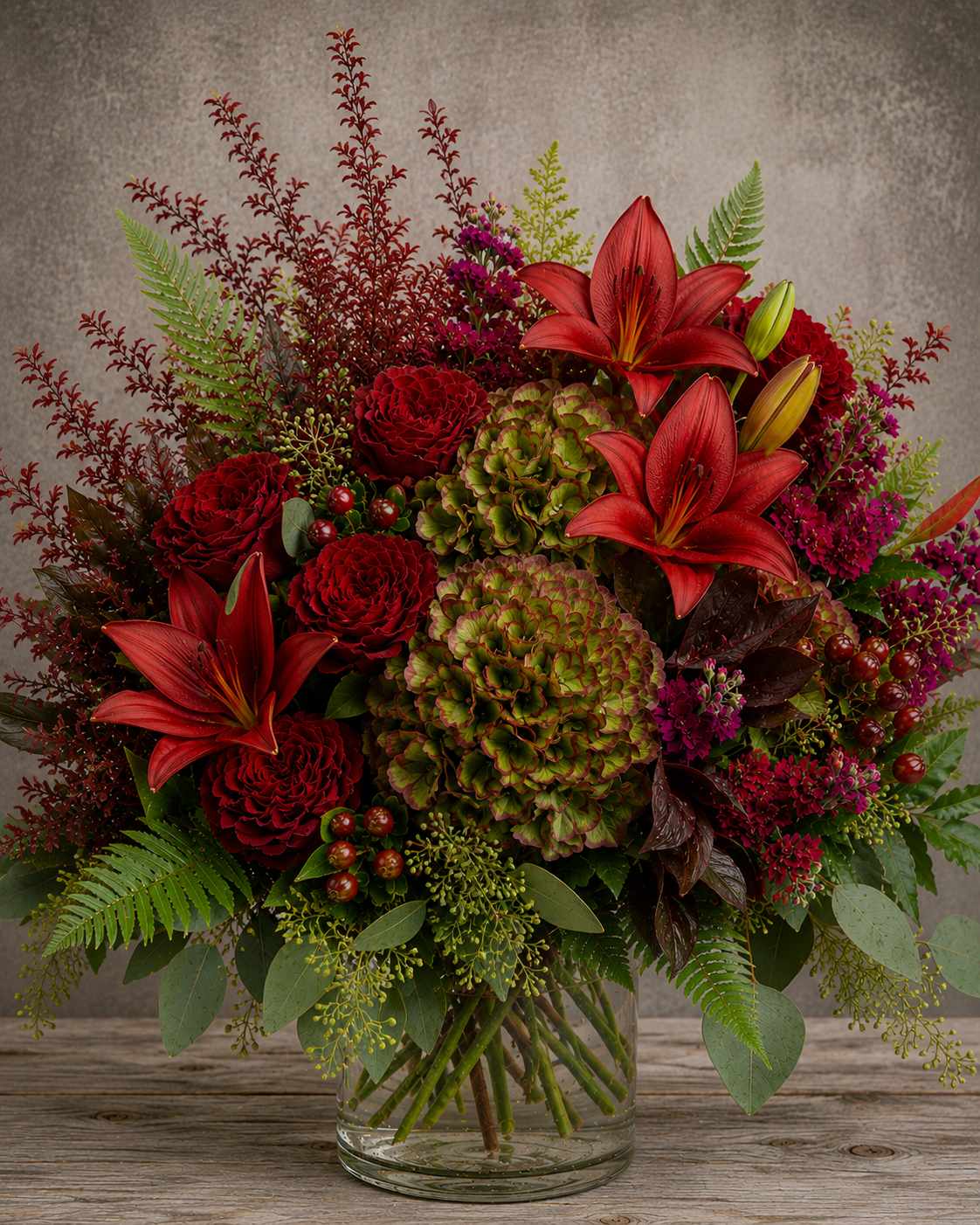 October Meadow fall floral arrangement with red roses, Antique green hydrangea, red lilies, burgundy stock flower, burgundy hypericum berries and textured greenery in a clear glass vase.