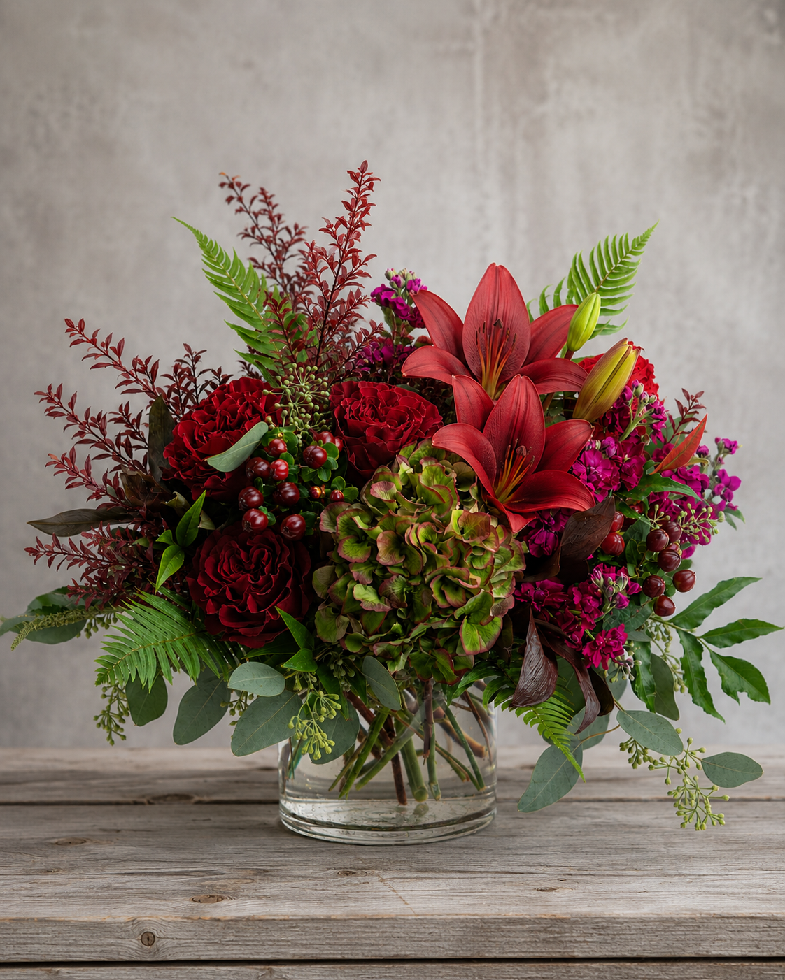 October Meadow fall floral arrangement with red roses, Antique green hydrangea, red lilies, burgundy stock flower, burgundy hypericum berries and textured greenery in a clear glass vase.