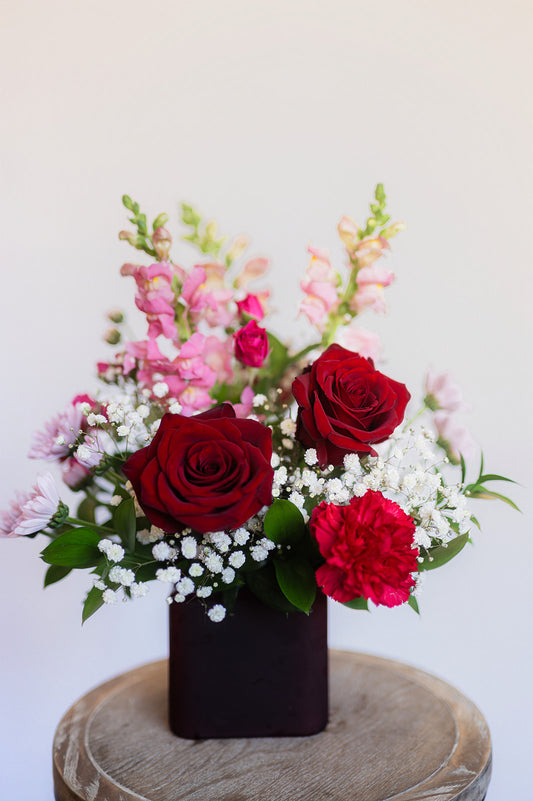 Bouquet of red and pink flowers in a dark vase on a wooden surface with a white background