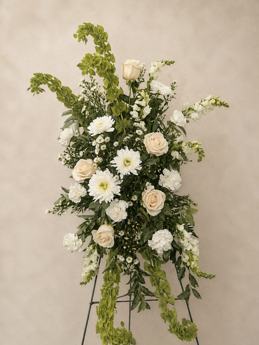 A large floral arrangement featuring white and cream roses, daisies, asters, and green foliage on a black metal stand, set against a plain beige background.